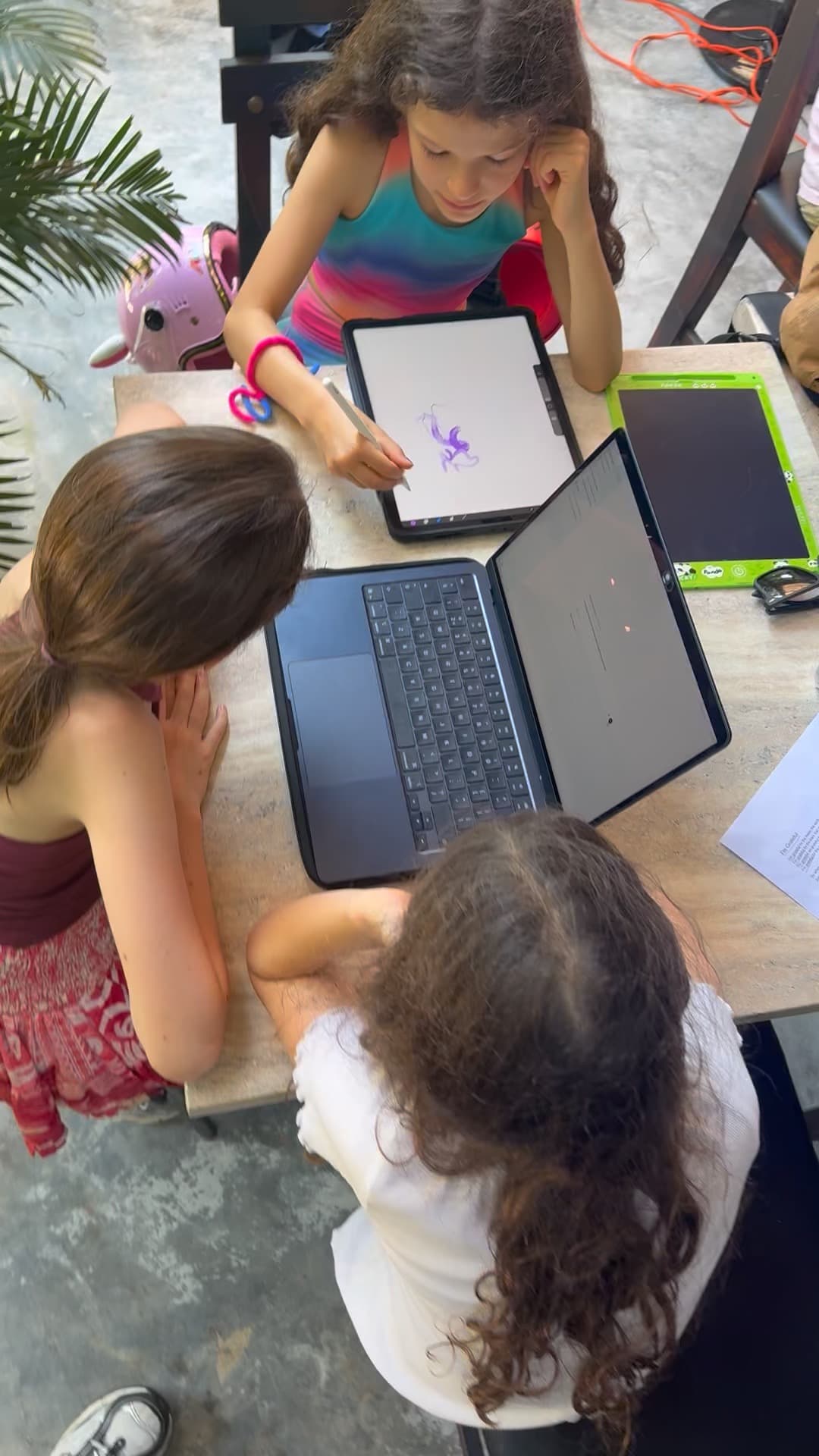 Three girls working together at a table, one drawing on a tablet with a stylus beside an open laptop