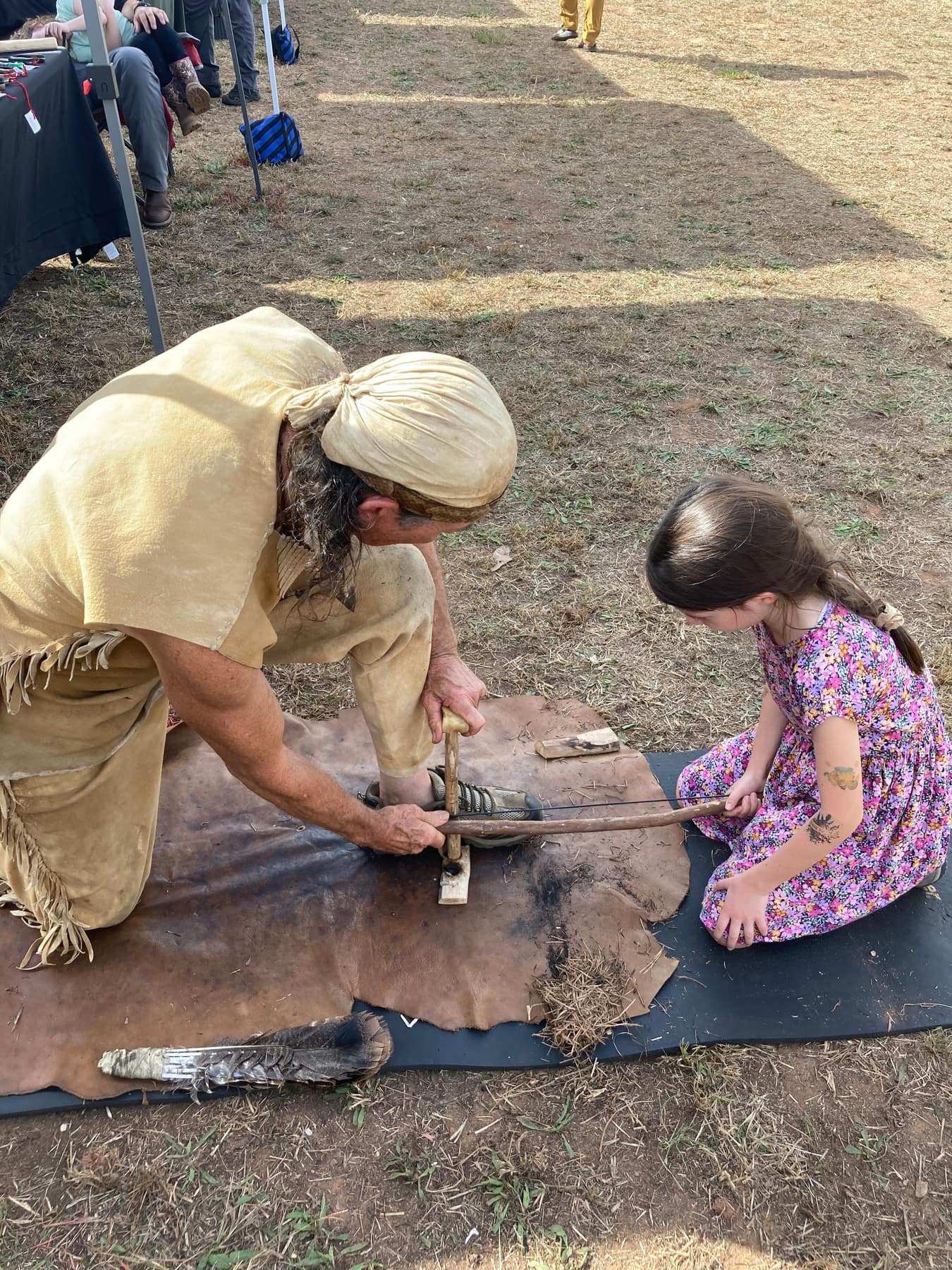 An instructor in traditional buckskin clothing showing a girl how to start a fire with a bow drill
