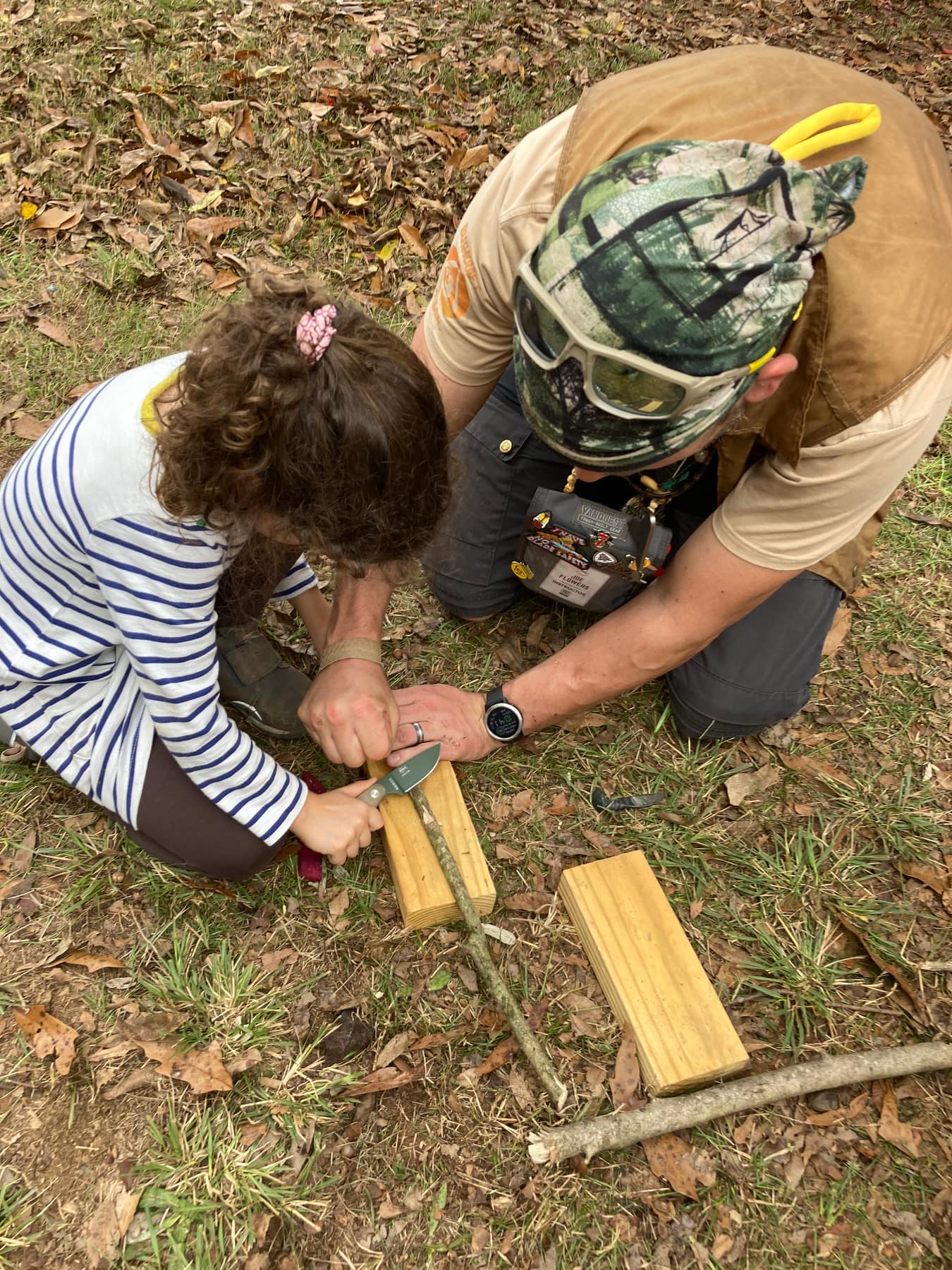 An instructor guiding a child carving wood with a knife