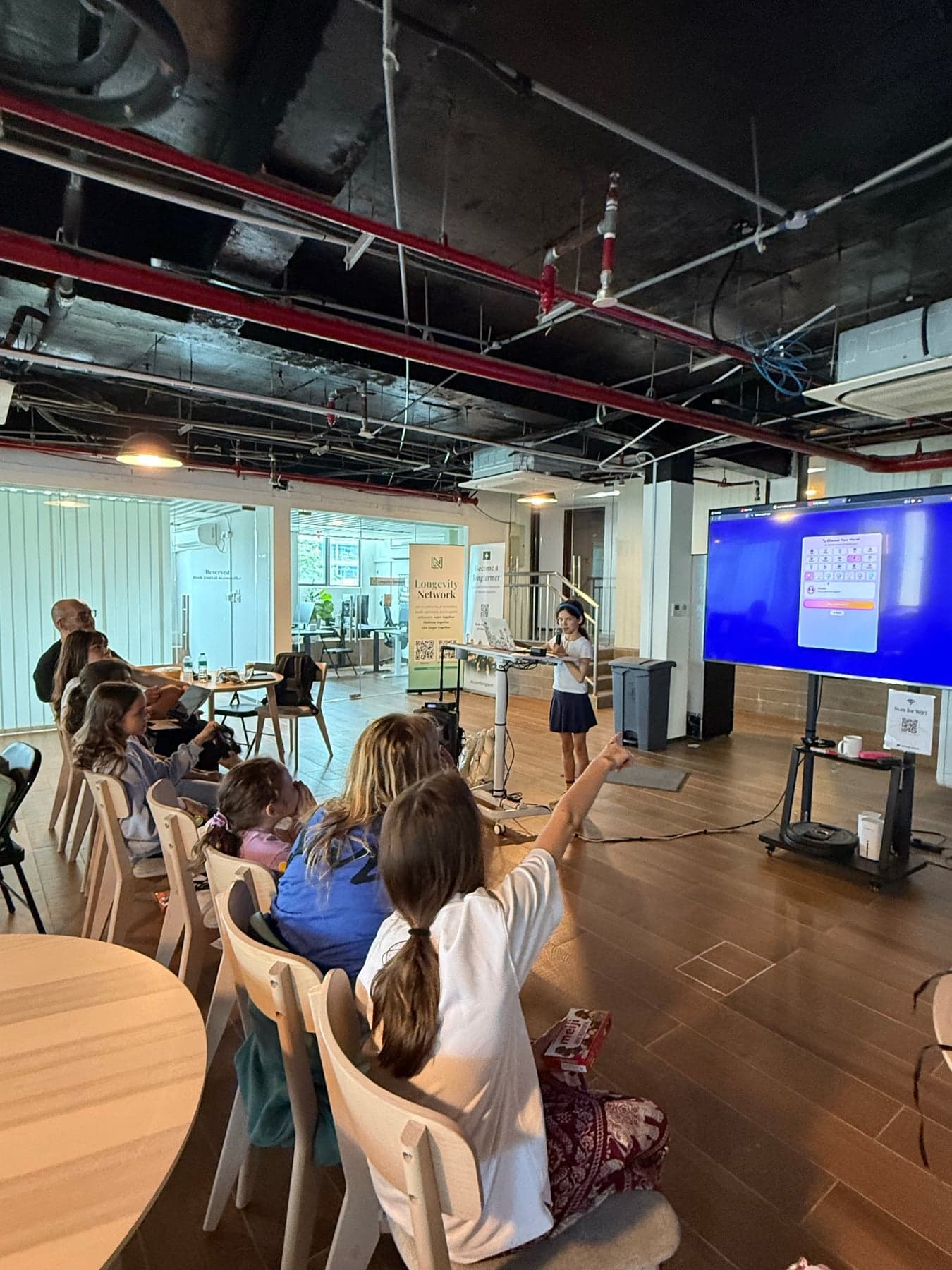 A young student presenting her app to a full room, screen glowing beside her