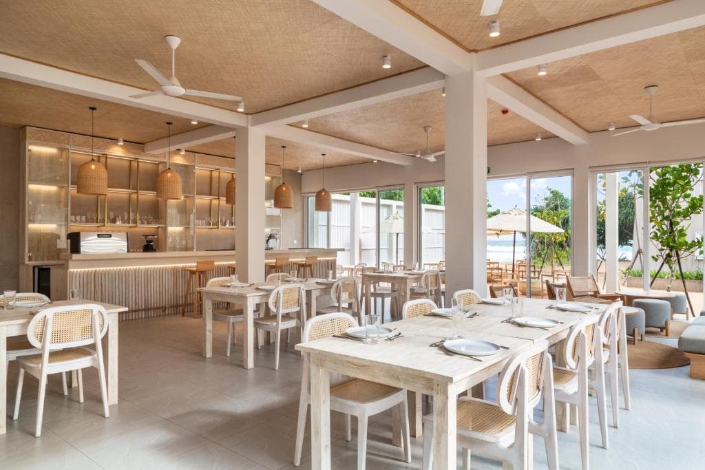 A bright cafe with whitewashed tables, cane chairs, pendant lamps, and the sea through the windows