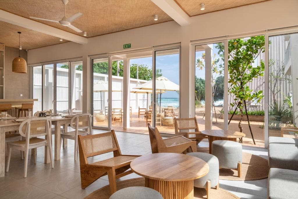 A lounge with cane armchairs and wooden tables, glass doors open to umbrellas and the sea