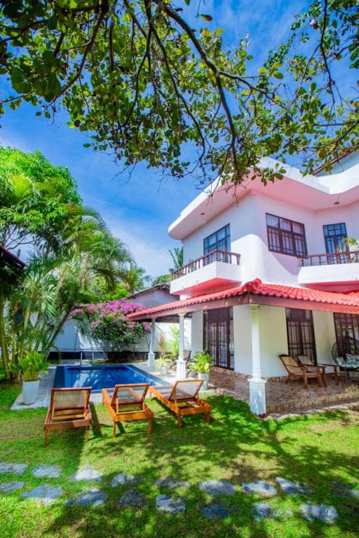 A whitewashed villa with red tile roofs, a pool, and wooden loungers in a green garden