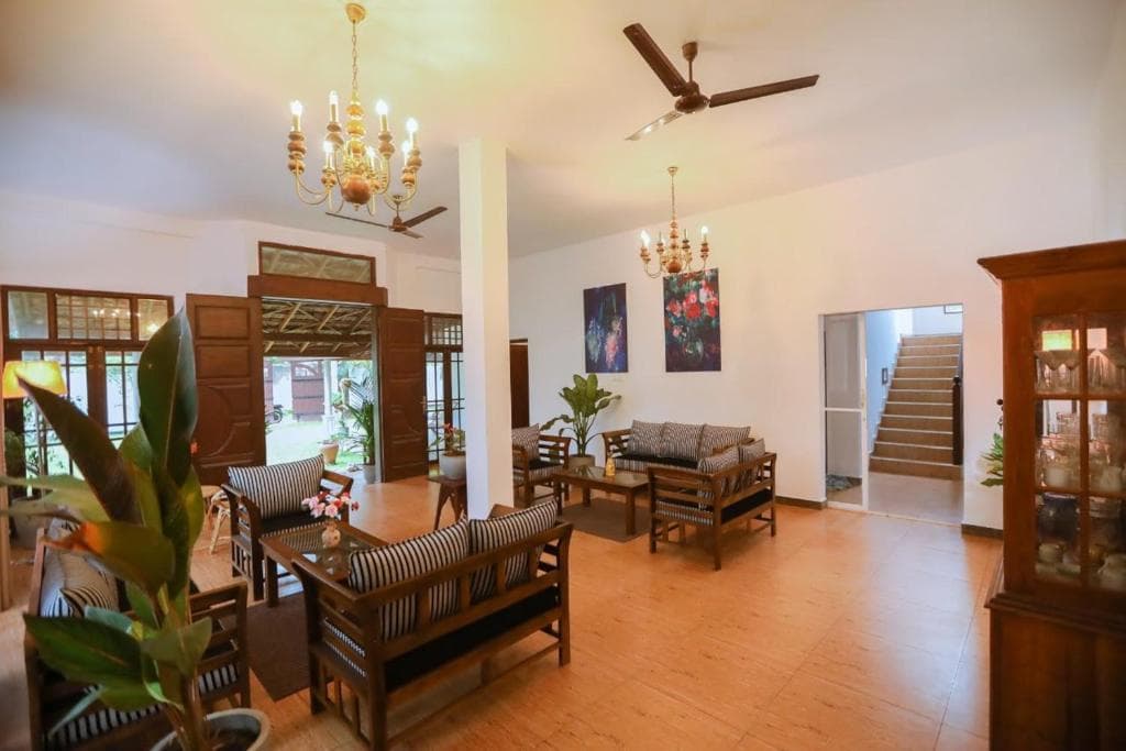 A bright shared lounge with wooden seating, chandeliers, and doors open toward the garden